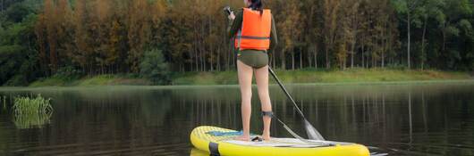 Young woman enjoying paddleboarding in mountain lake wearing short wetsuit and an orange life jacket.